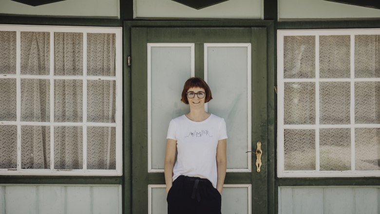 A woman stands in front of a green door with windows and lace curtains.