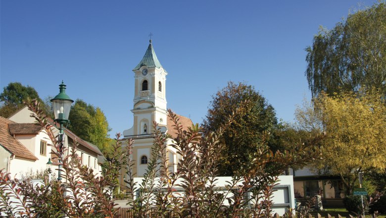 Walpersbach parish church with tower and surrounding buildings, surrounded by trees and bushes under a blue sky.