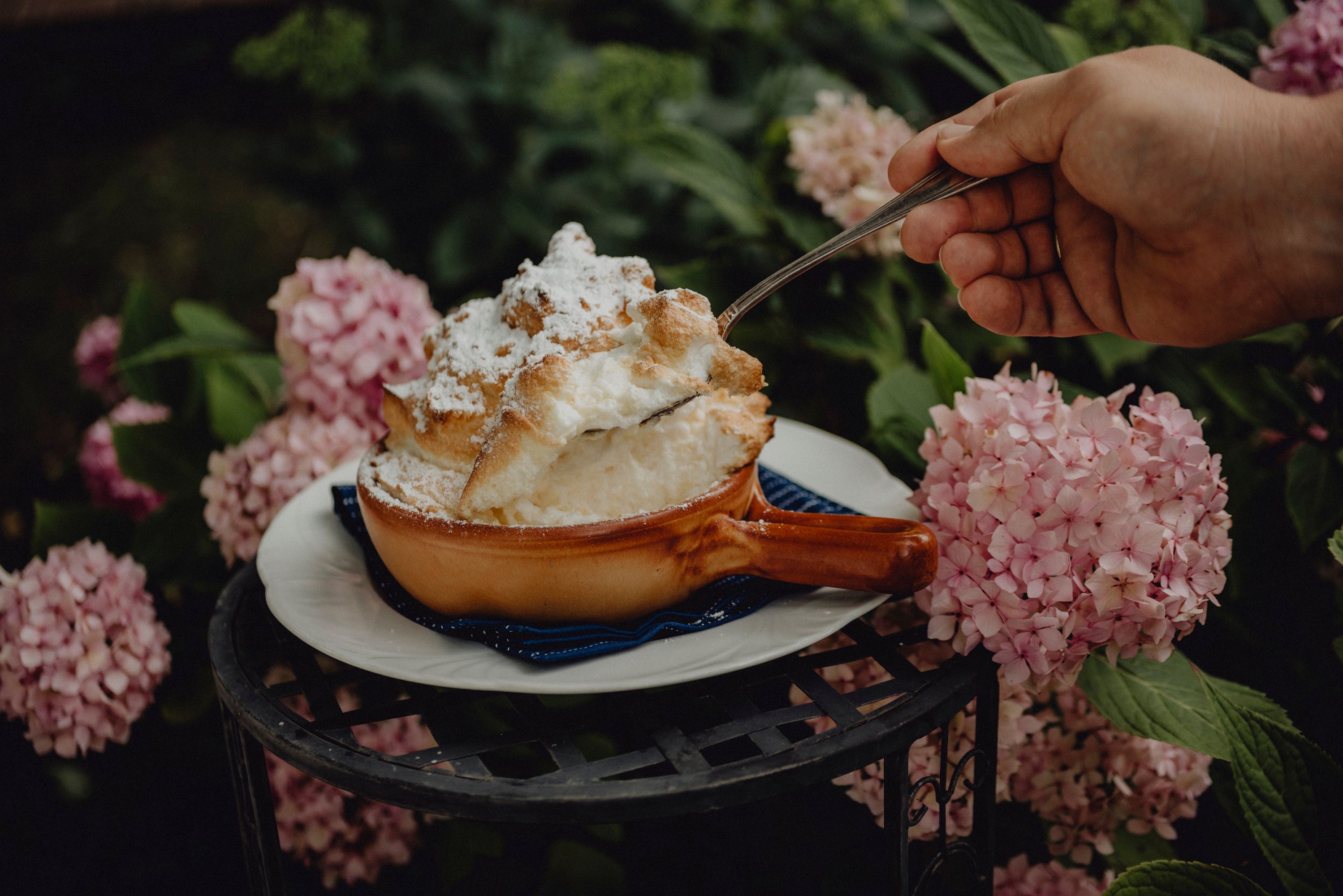A soufflé in a brown bowl, surrounded by pink hydrangeas, is served with a spoon.