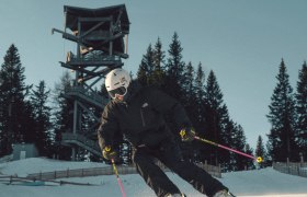 Skier in black clothing skiing on a groomed slope on the Semmering Hirschenkogel, with a lookout tower and trees in the background.