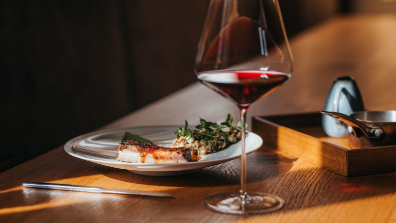 A plate of lamb belly, lettuce and wasabi cream next to a glass of red wine on a wooden table.