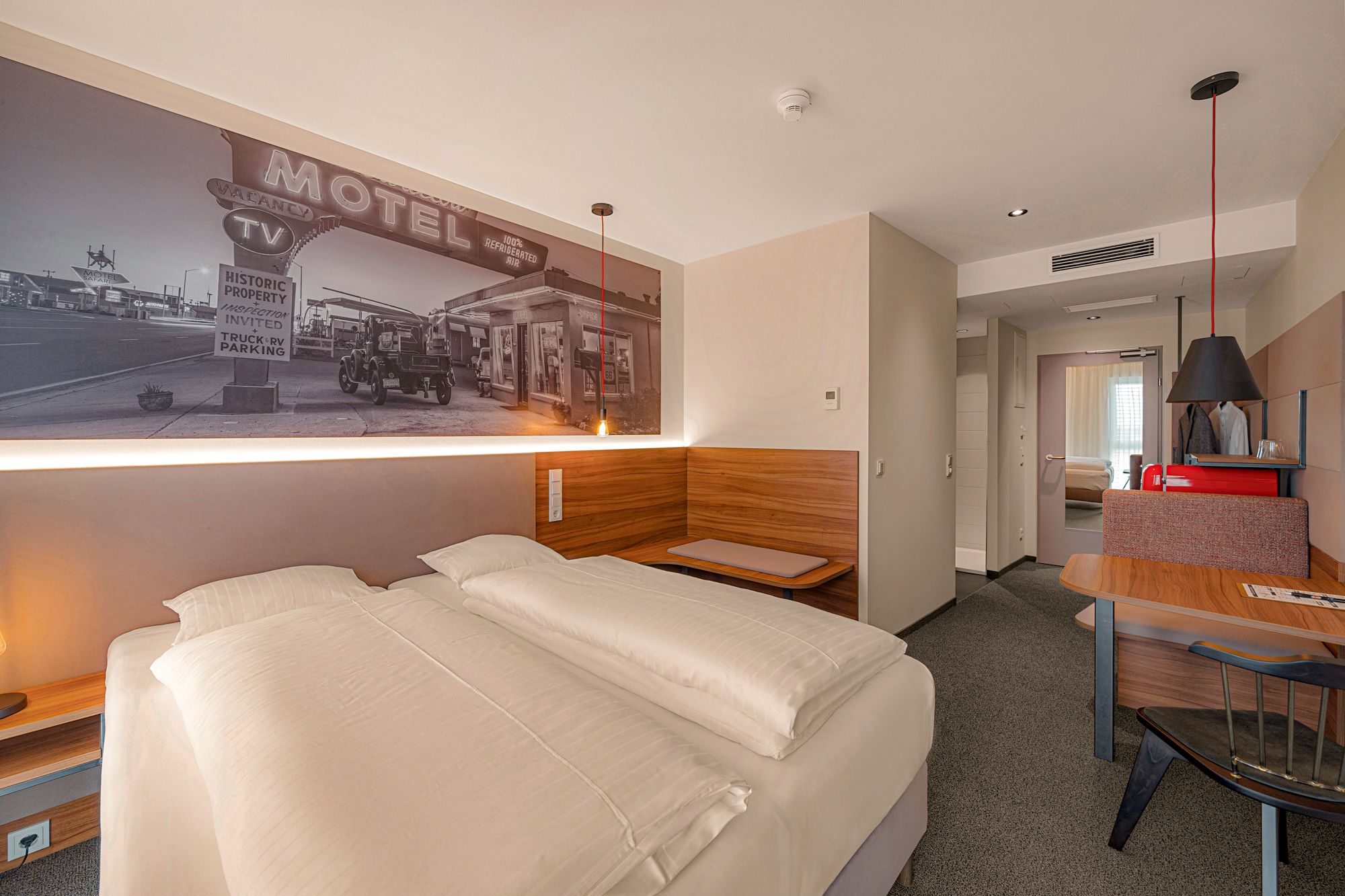 Modern hotel room with double bed, mural of an old motel, wooden furniture and desk.