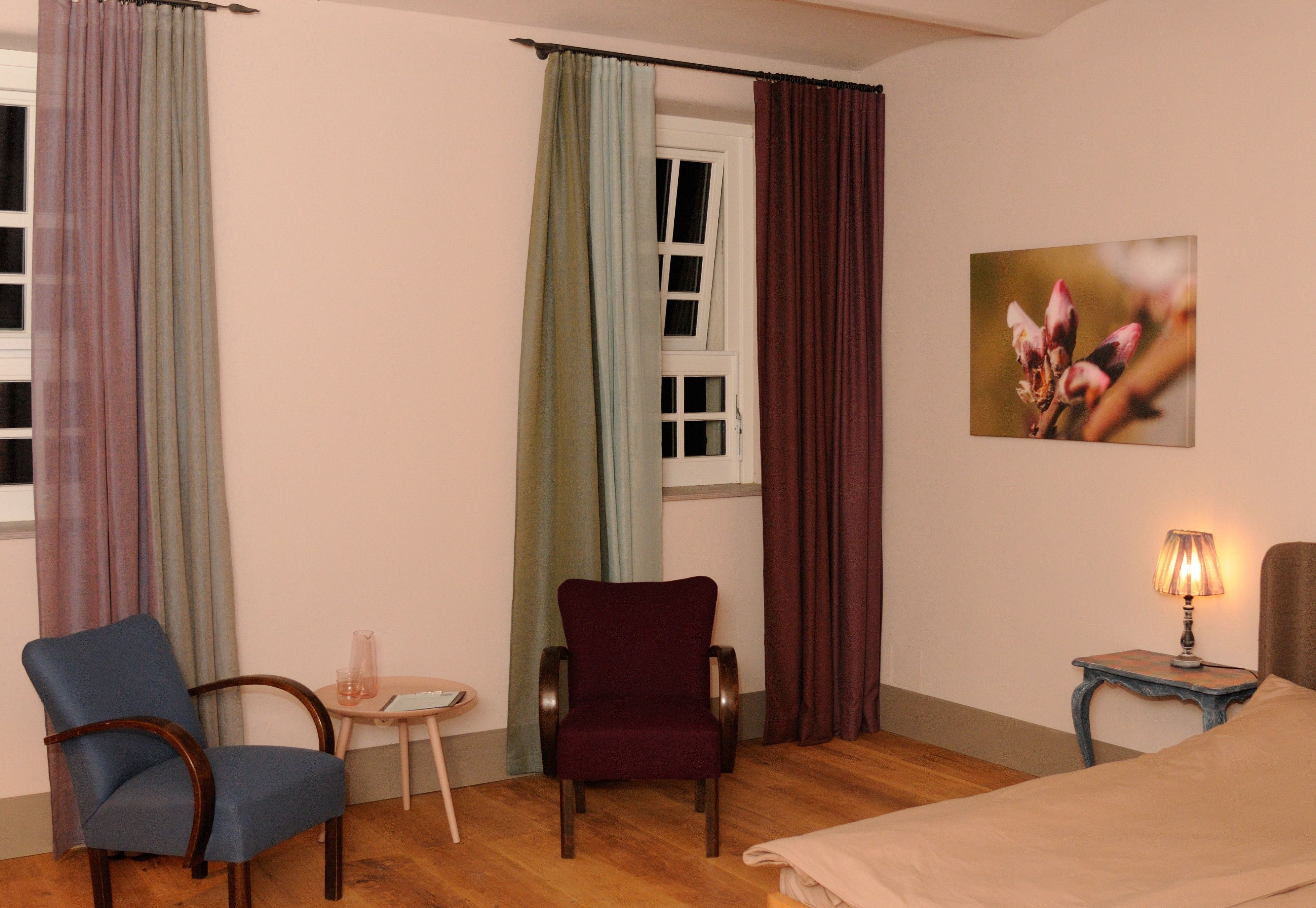 A cozy room with two armchairs, a small table, a bed and a lamp. A picture with a flower motif hangs on the wall.