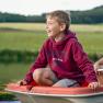 A boy in a red hoodie sits smiling on a boat on a pond.