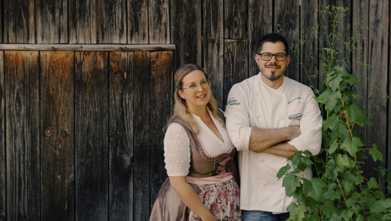 A couple is standing in front of a wooden wall, she is wearing a dirndl, he a chef's jacket.