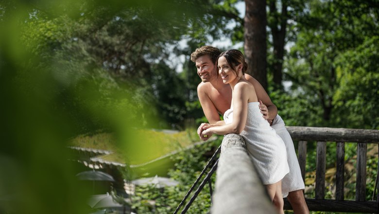 A couple in towels lean smiling against a railing outside, surrounded by green trees.