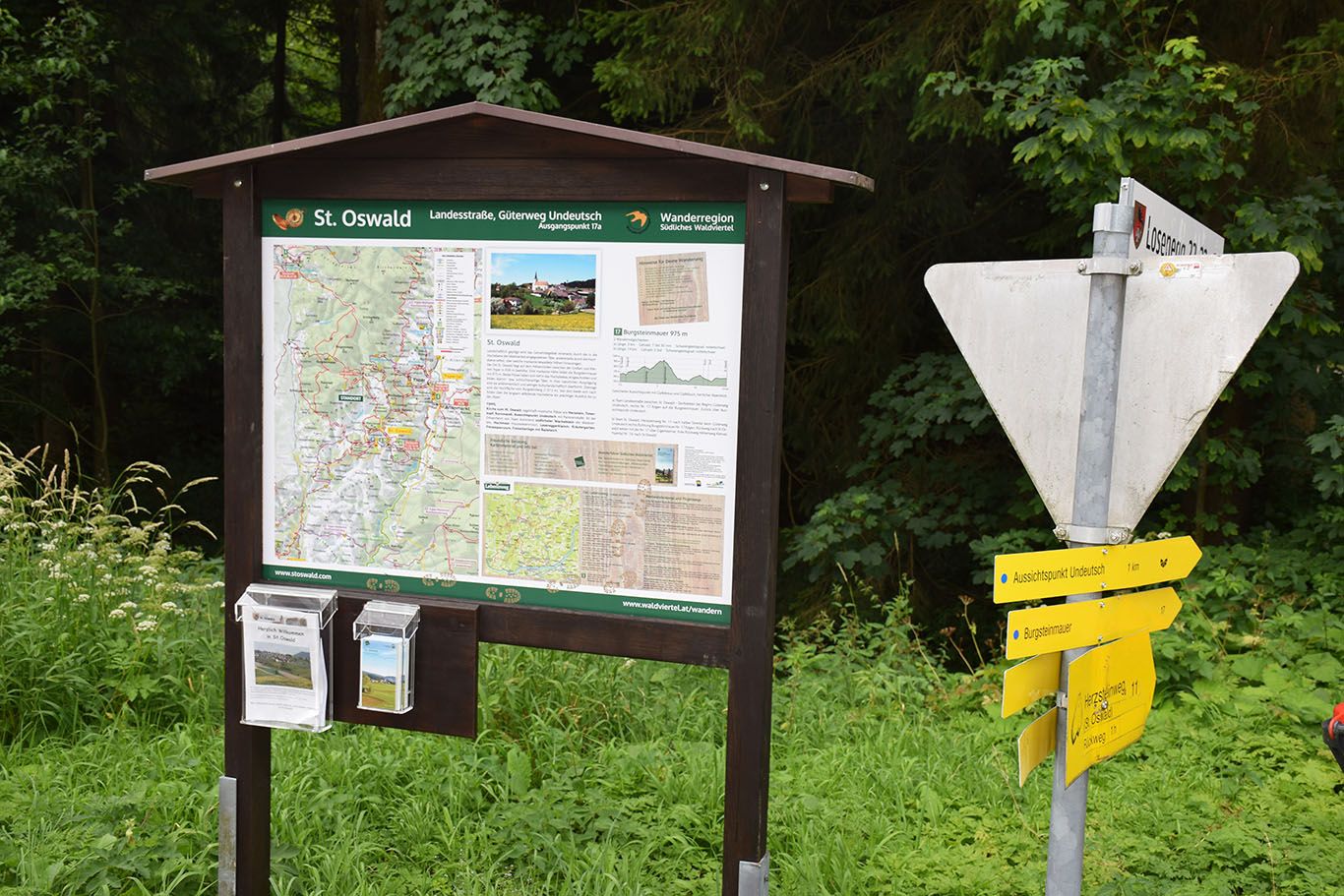 Hiking signpost and information board in St. Oswald, surrounded by forest and greenery.