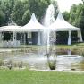 A wedding pavilion and tents next to a pond with a fountain in a garden.