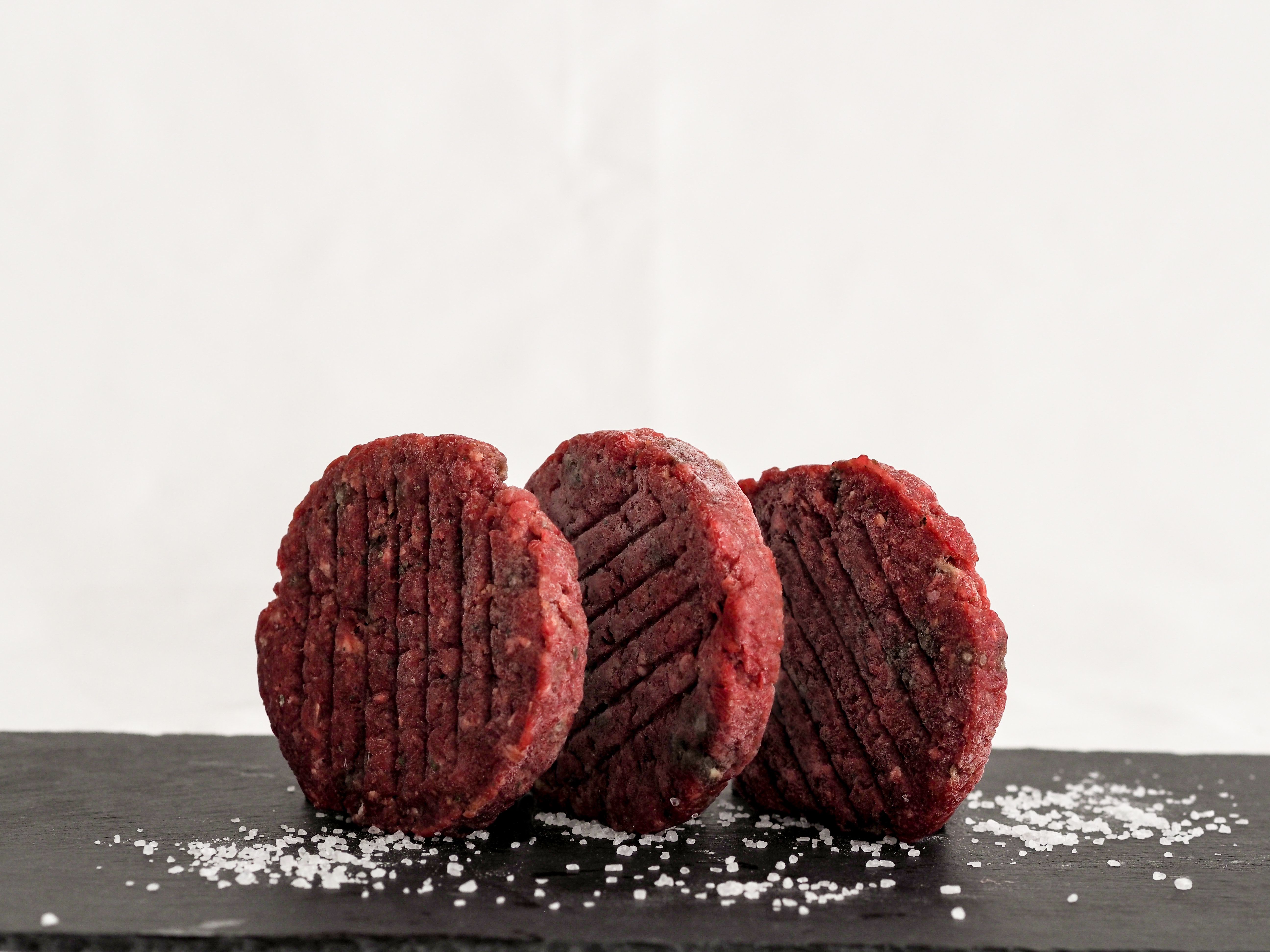 Three raw burger patties are placed upright on a dark slate plate, with salt next to them.