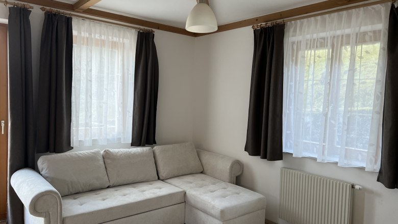 A living room with a beige corner sofa, two windows with dark curtains and a radiator underneath.