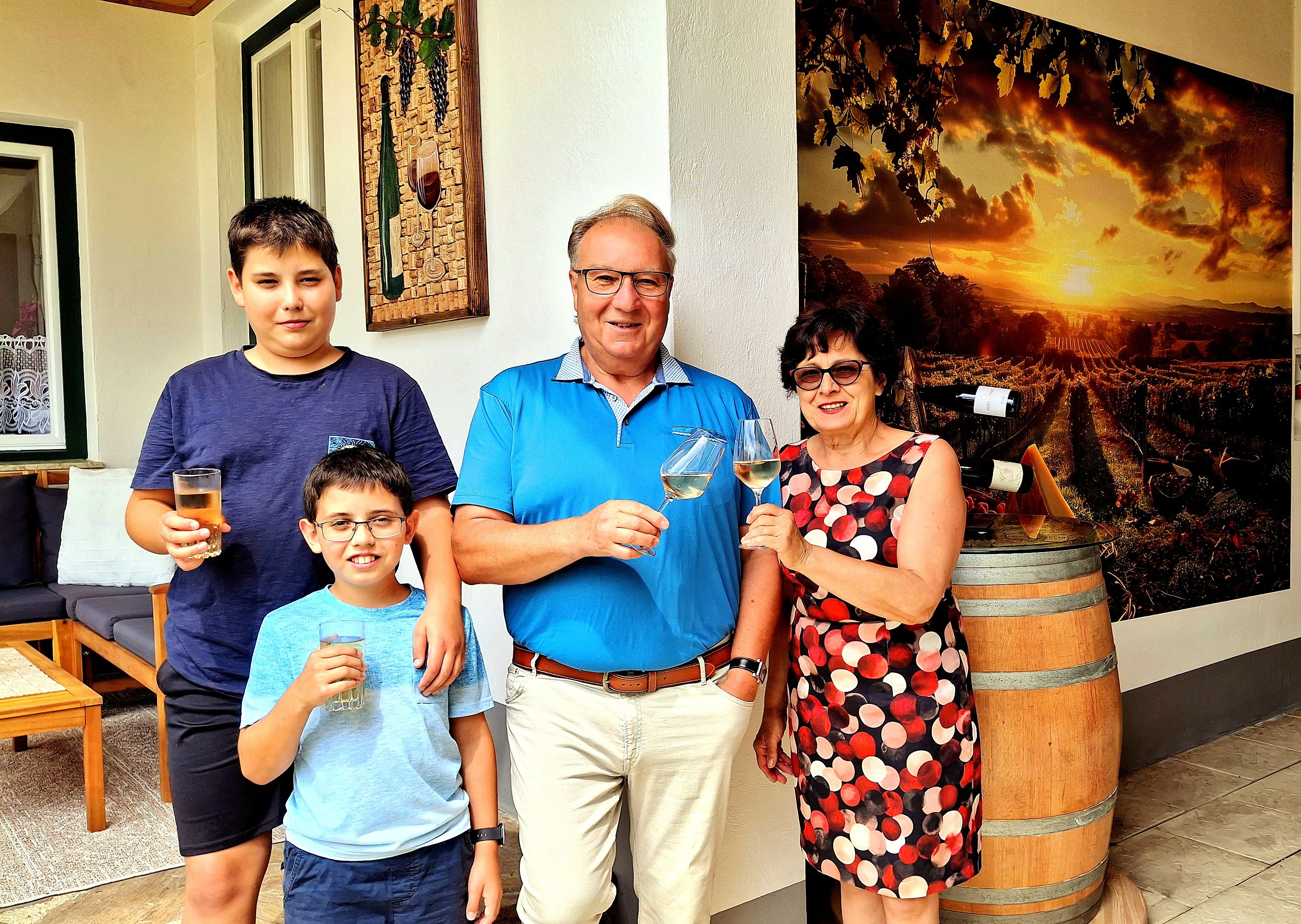 A family stands in front of a wine barrel and a picture of vineyards, holding glasses and smiling at the camera.