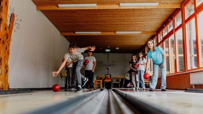 Children play skittles in a room with wooden walls and large windows.