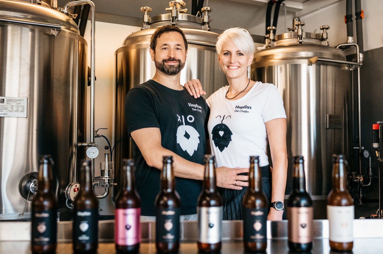 A man and a woman stand smiling in front of brewing tanks, with various bottles of beer in the foreground.