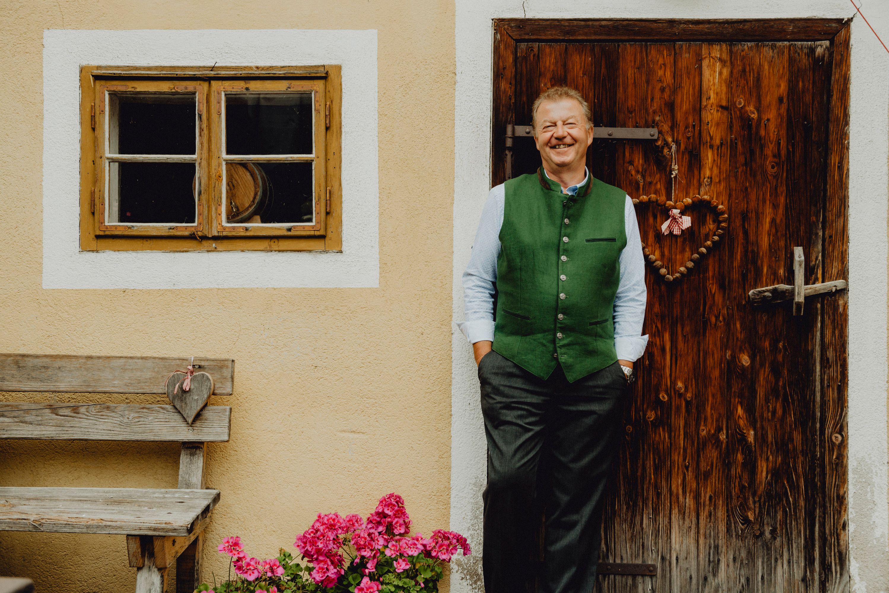 A man in traditional dress stands smiling in front of a wooden door next to a window and a bench with flowers.
