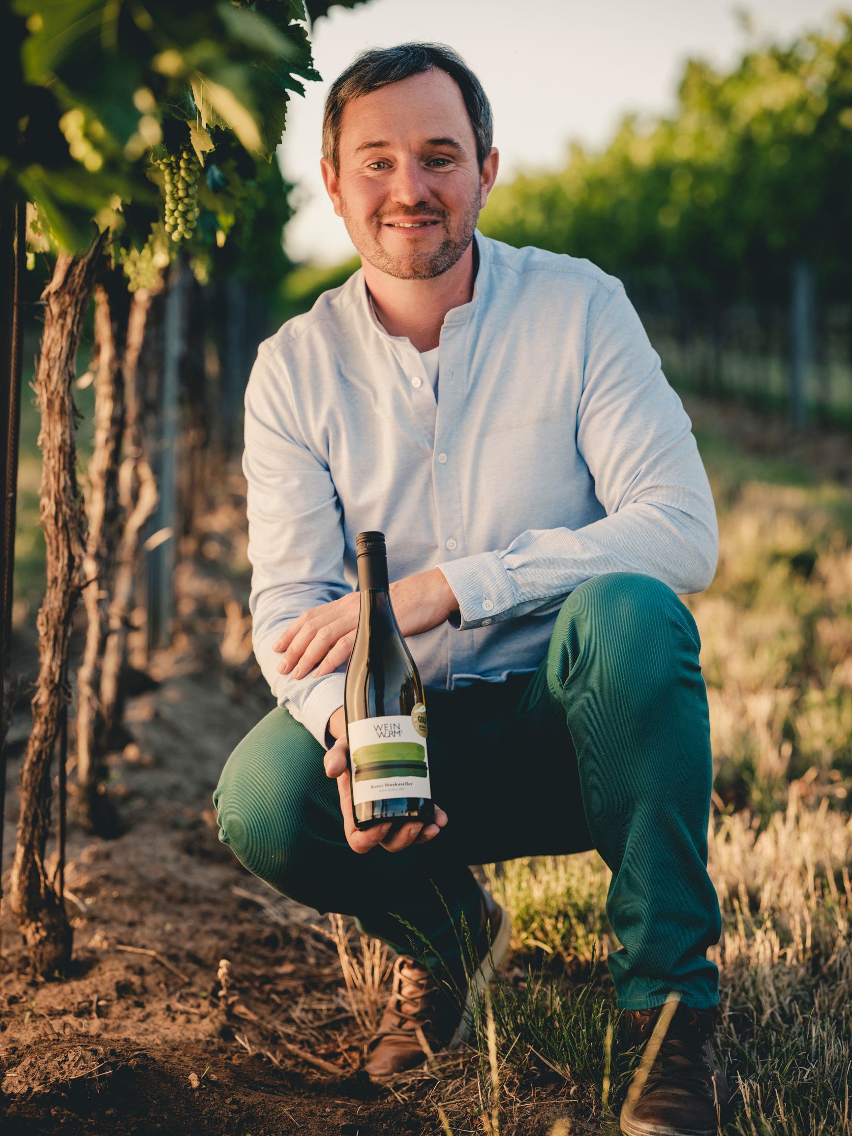 A man kneels in a vineyard and holds a bottle of wine in his hand.