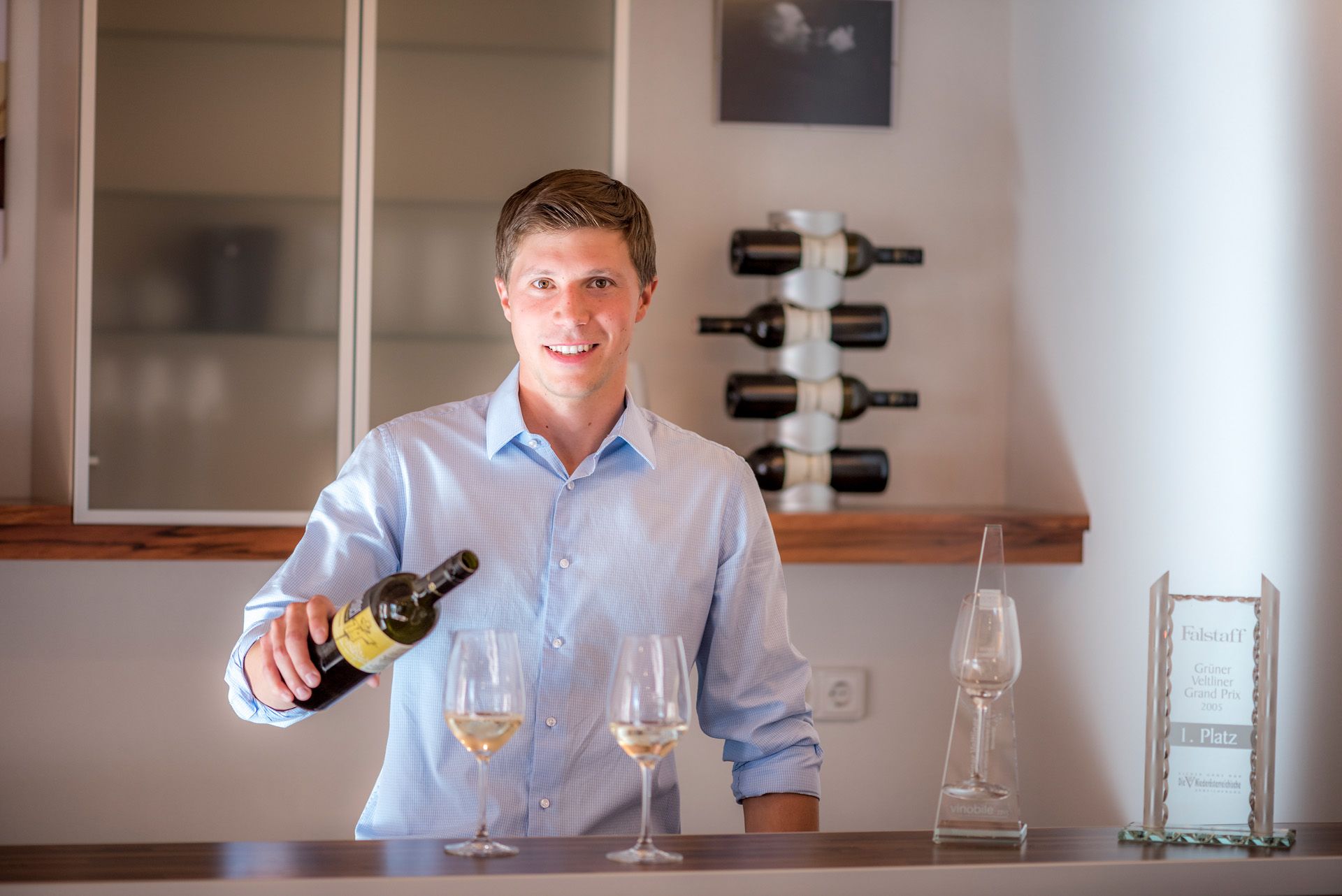 A young man in a blue shirt pours wine into a glass, with bottles of wine and an award in the background.