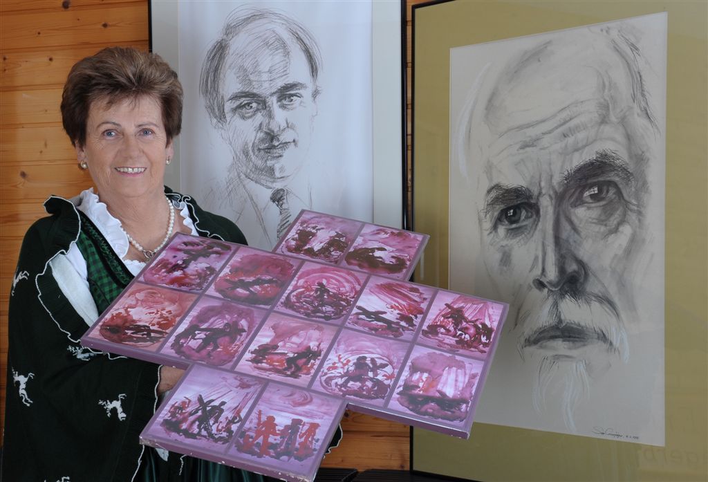 Woman in traditional costume holding artwork in front of two portraits.