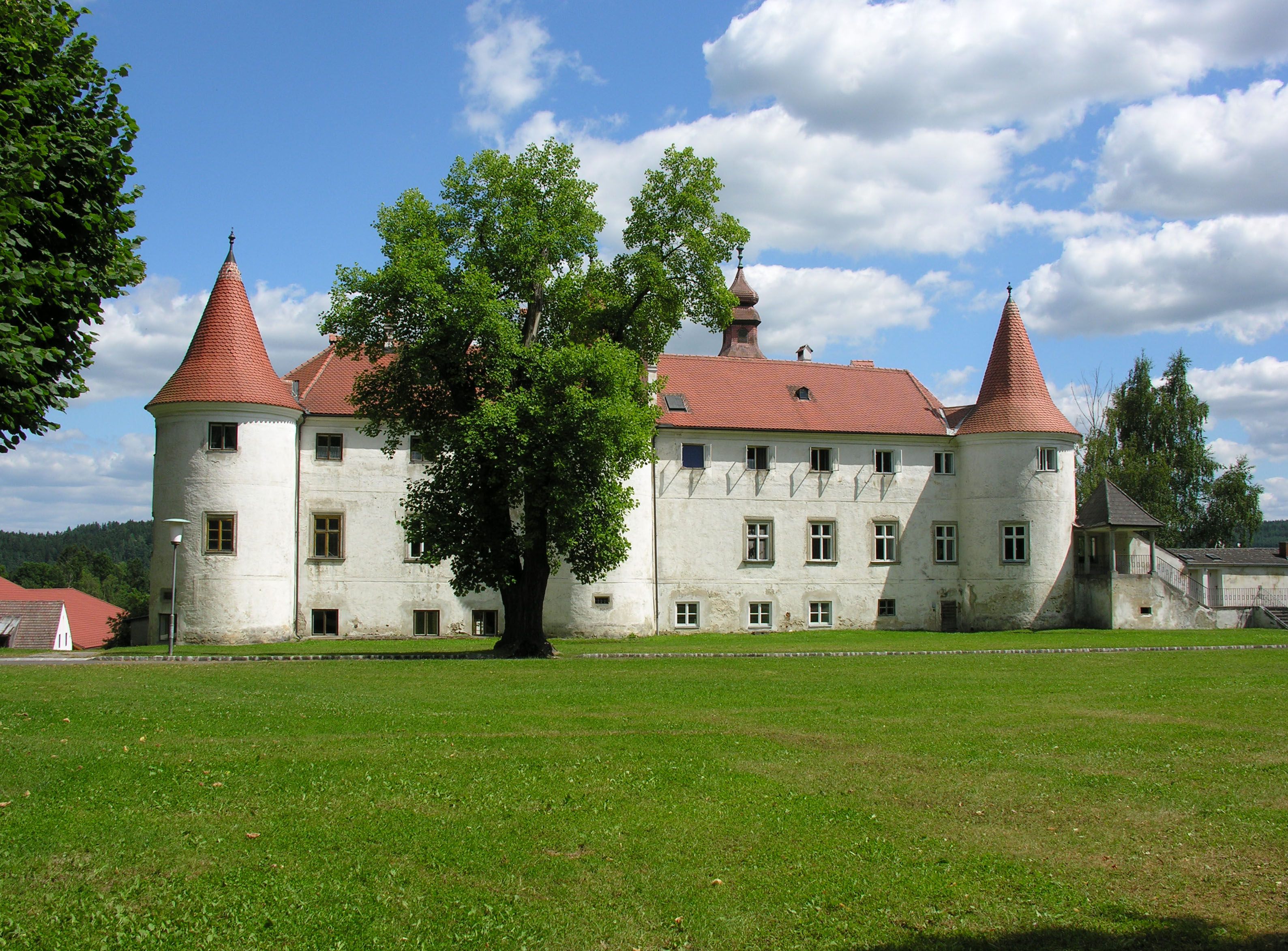 A historic castle with red roofs and towers, surrounded by green meadows and trees, under a blue sky with white clouds.