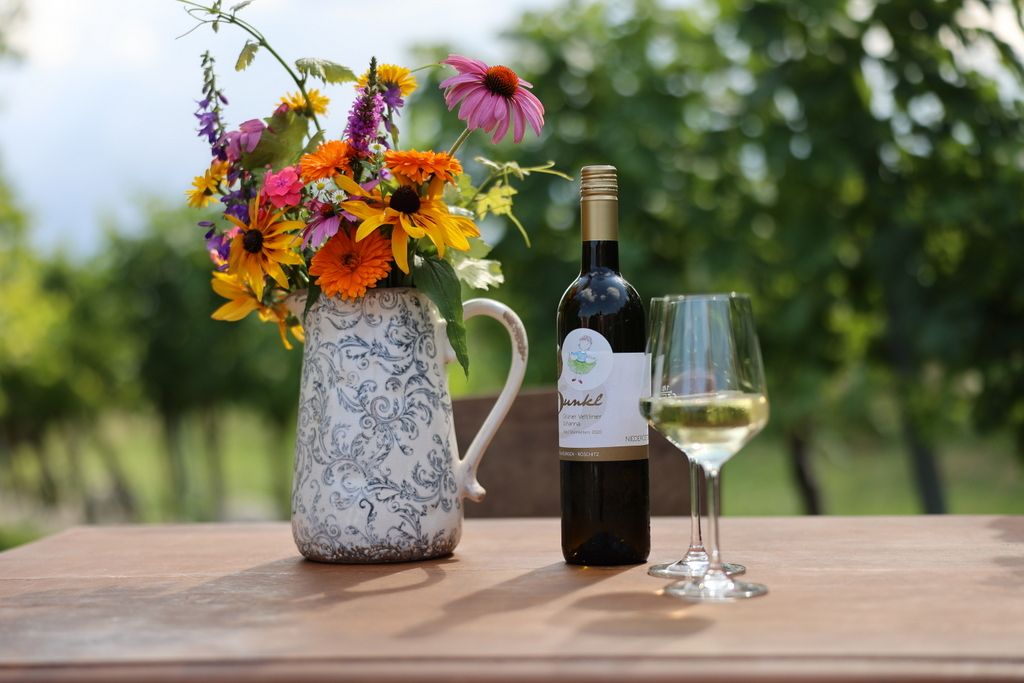 Wine bottle and glass on table with bouquet of flowers in vase, grapevines in background.