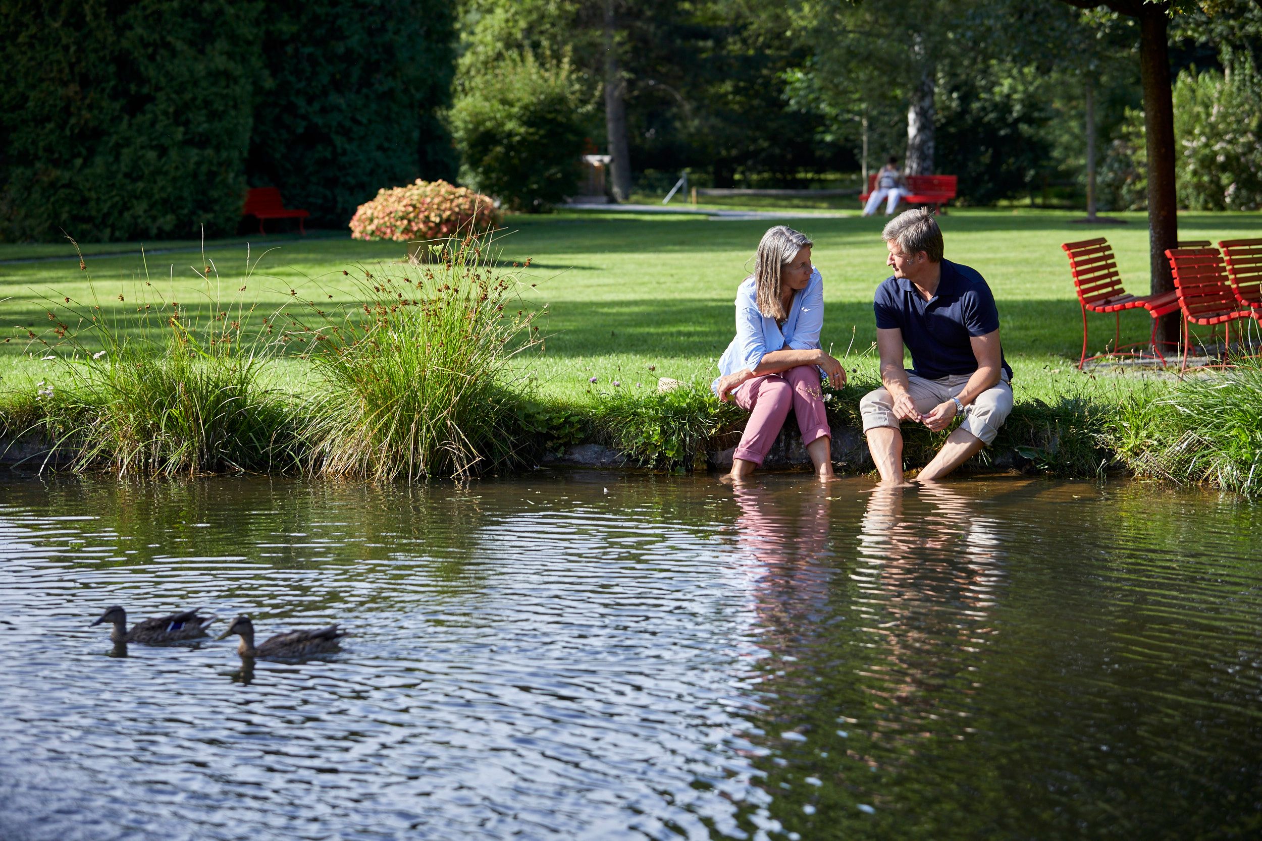 A couple sits on the bank of a pond in the spa gardens of Bad Schönau, their feet in the water. Two ducks swim in the foreground.