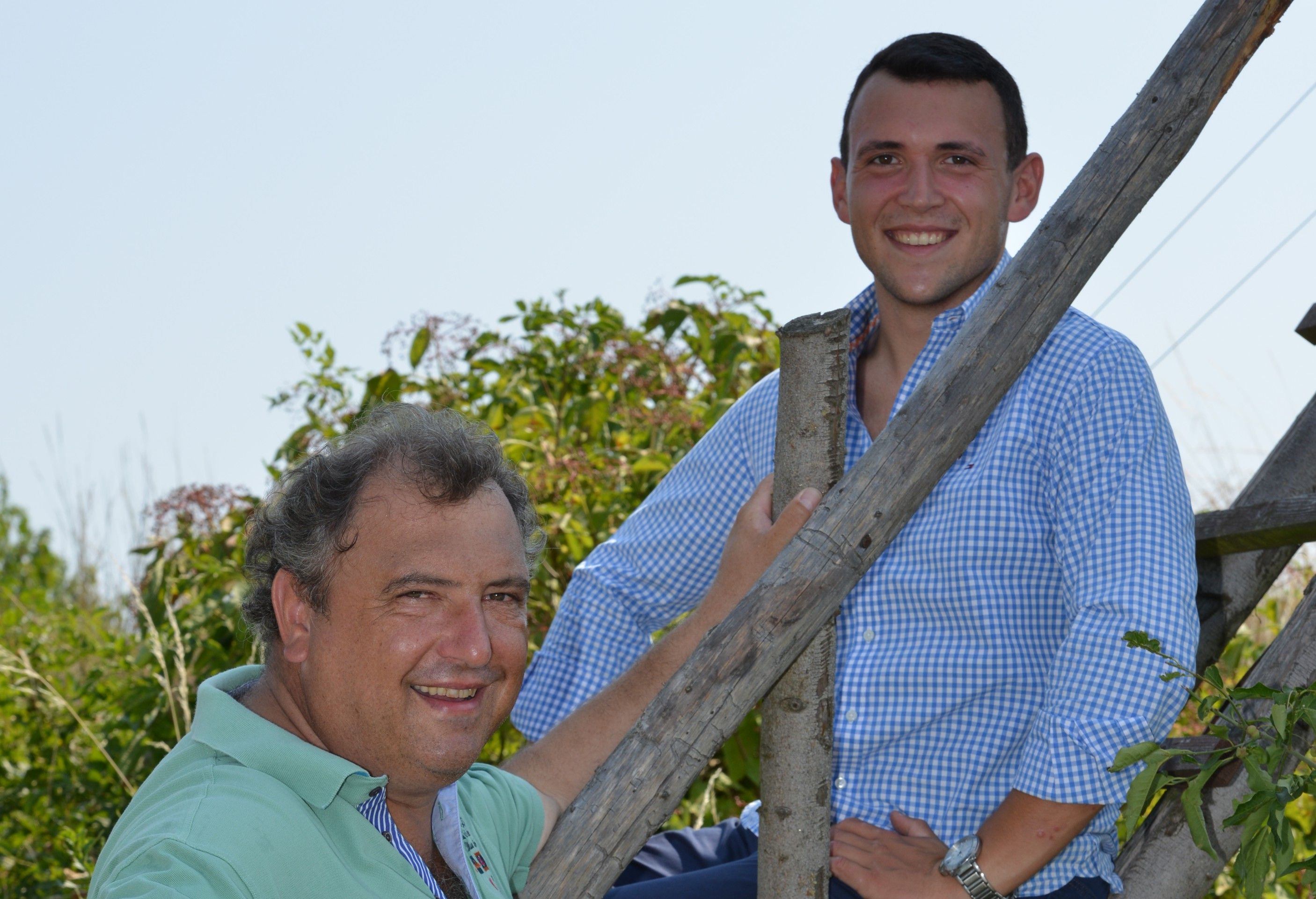 Two men smile into the camera, one sitting, the other standing on a wooden ladder outside.
