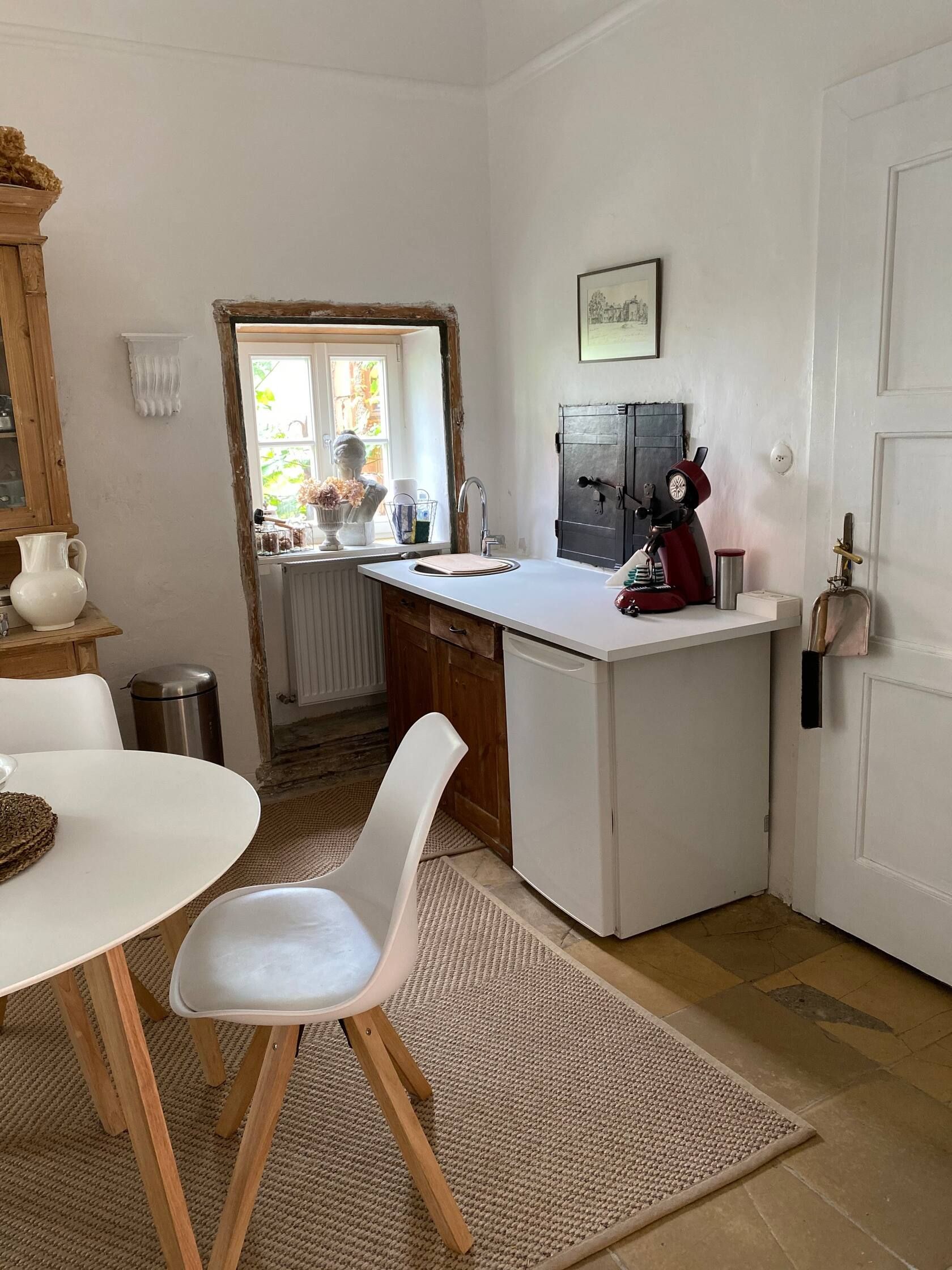 Kitchen area with small table, chair, sink and coffee machine. Windows with decoration and bright walls.