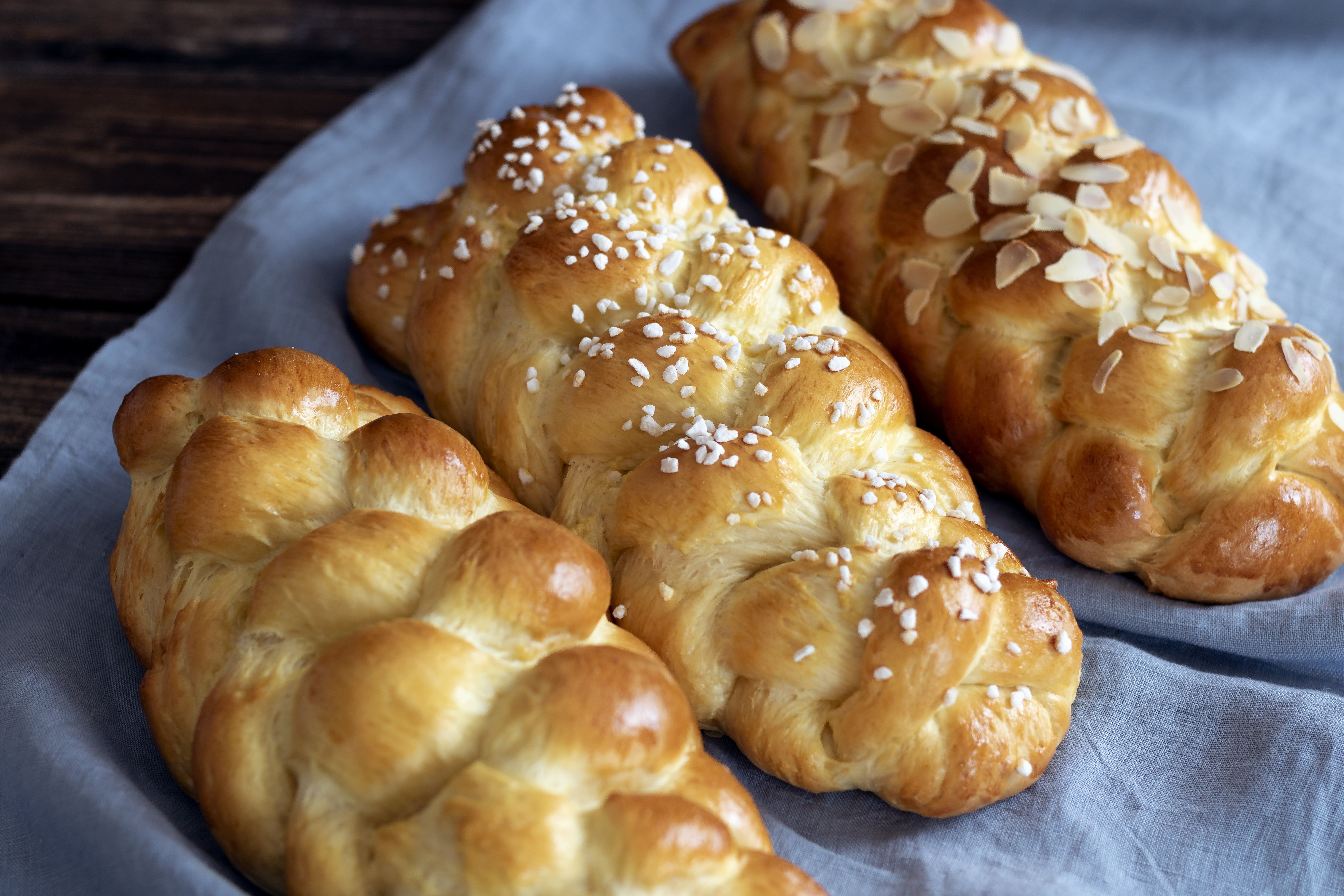 Three braided brioche striezel on a blue cloth, one sprinkled with almonds, one with sugar crystals.