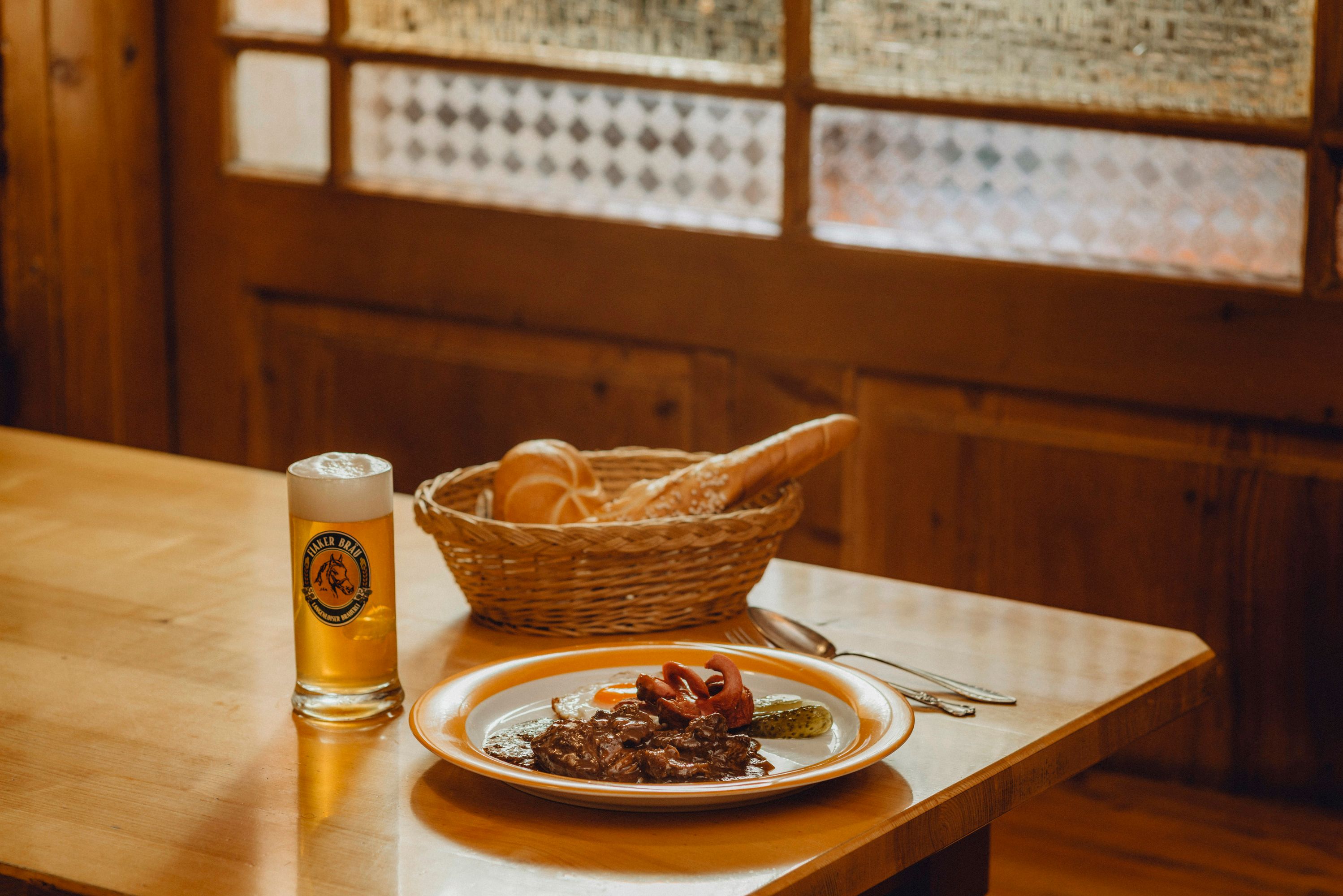 A plate with Fiaker goulash, fried egg and gherkin, next to it a glass of beer and a basket of bread on a wooden table.