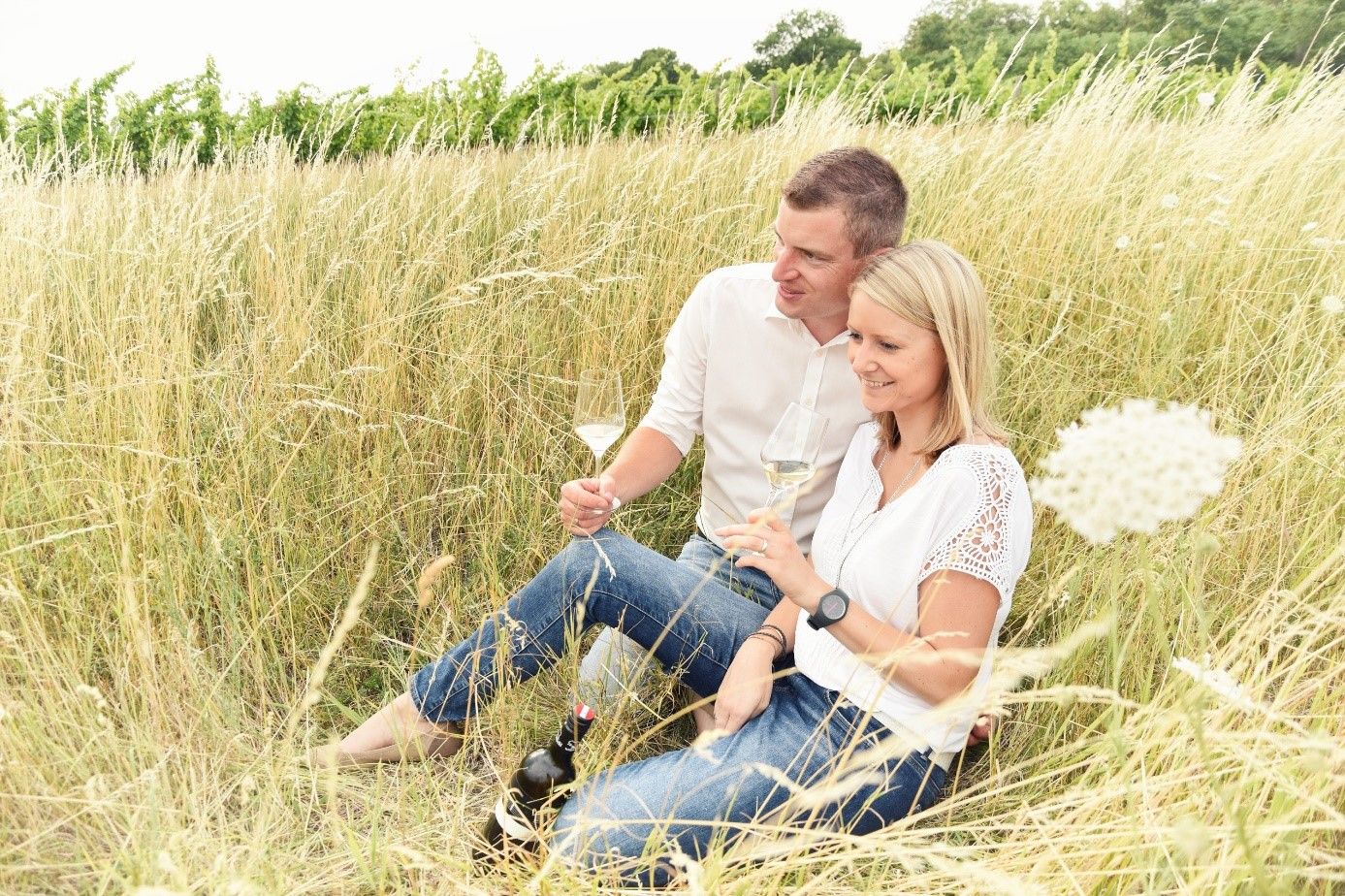 A couple sits in the tall grass and drinks wine.