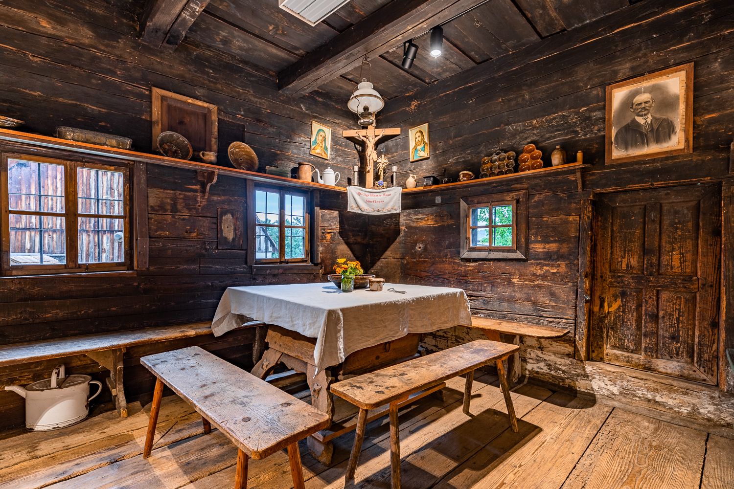 Rustic room with wooden walls, table with white cloth, benches, religious pictures and antique objects.