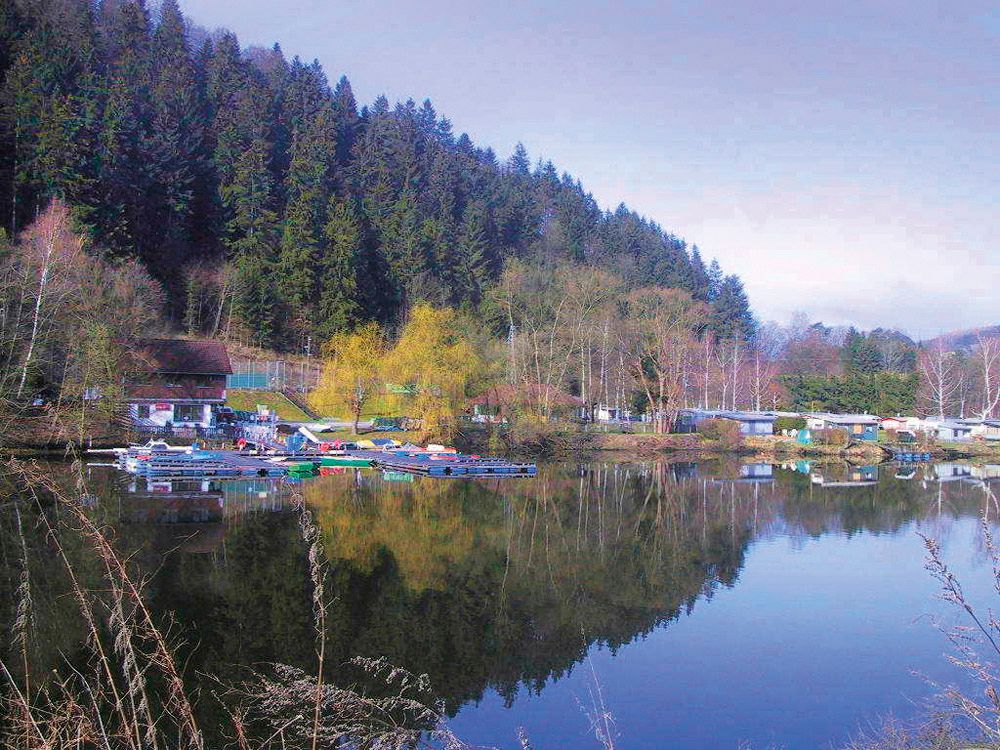 Lakeside campsite with caravans and boats on the shore of a lake, surrounded by forest.
