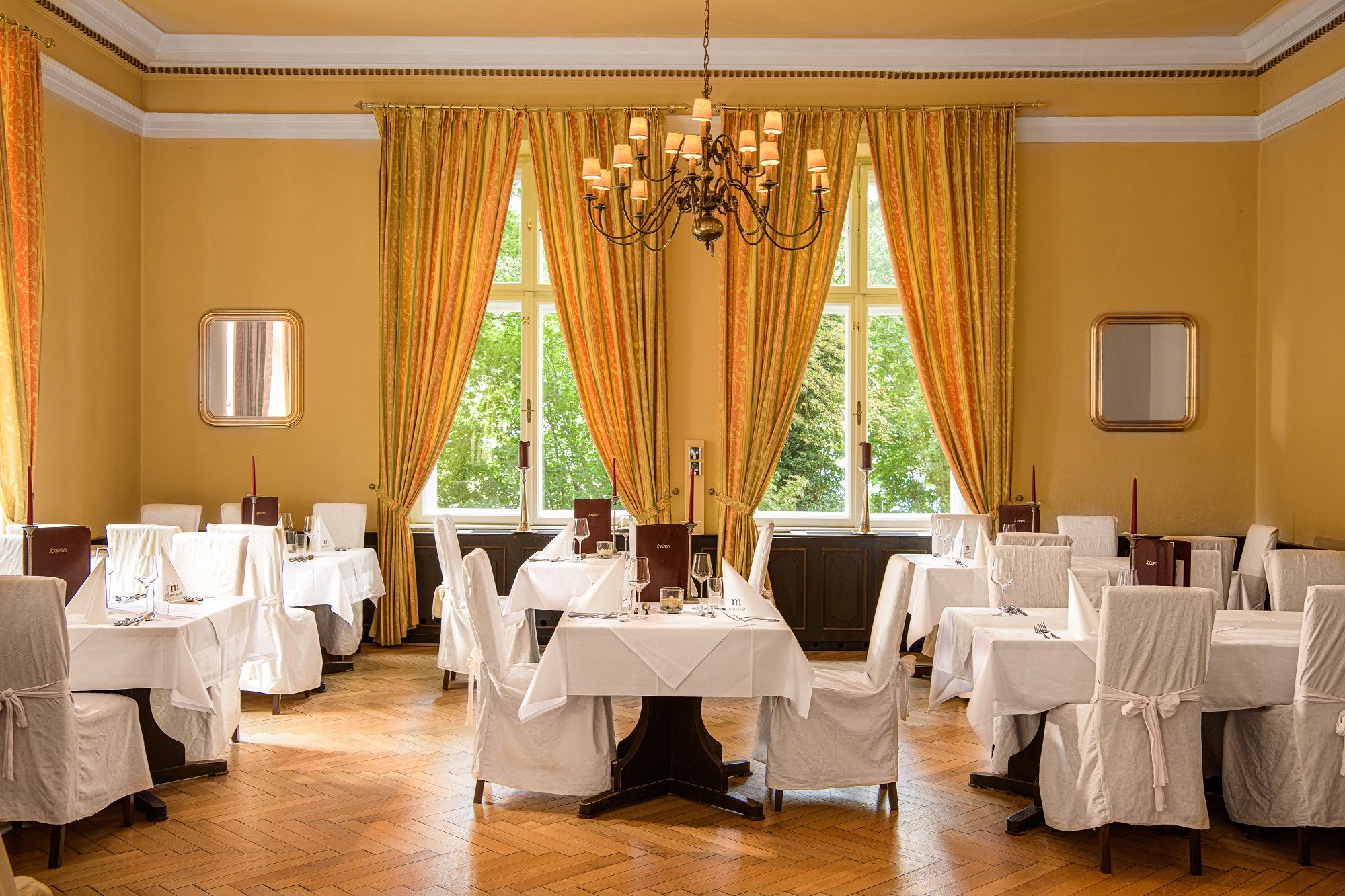 Elegant hotel restaurant with laid tables, white chair covers and large windows with yellow curtains.