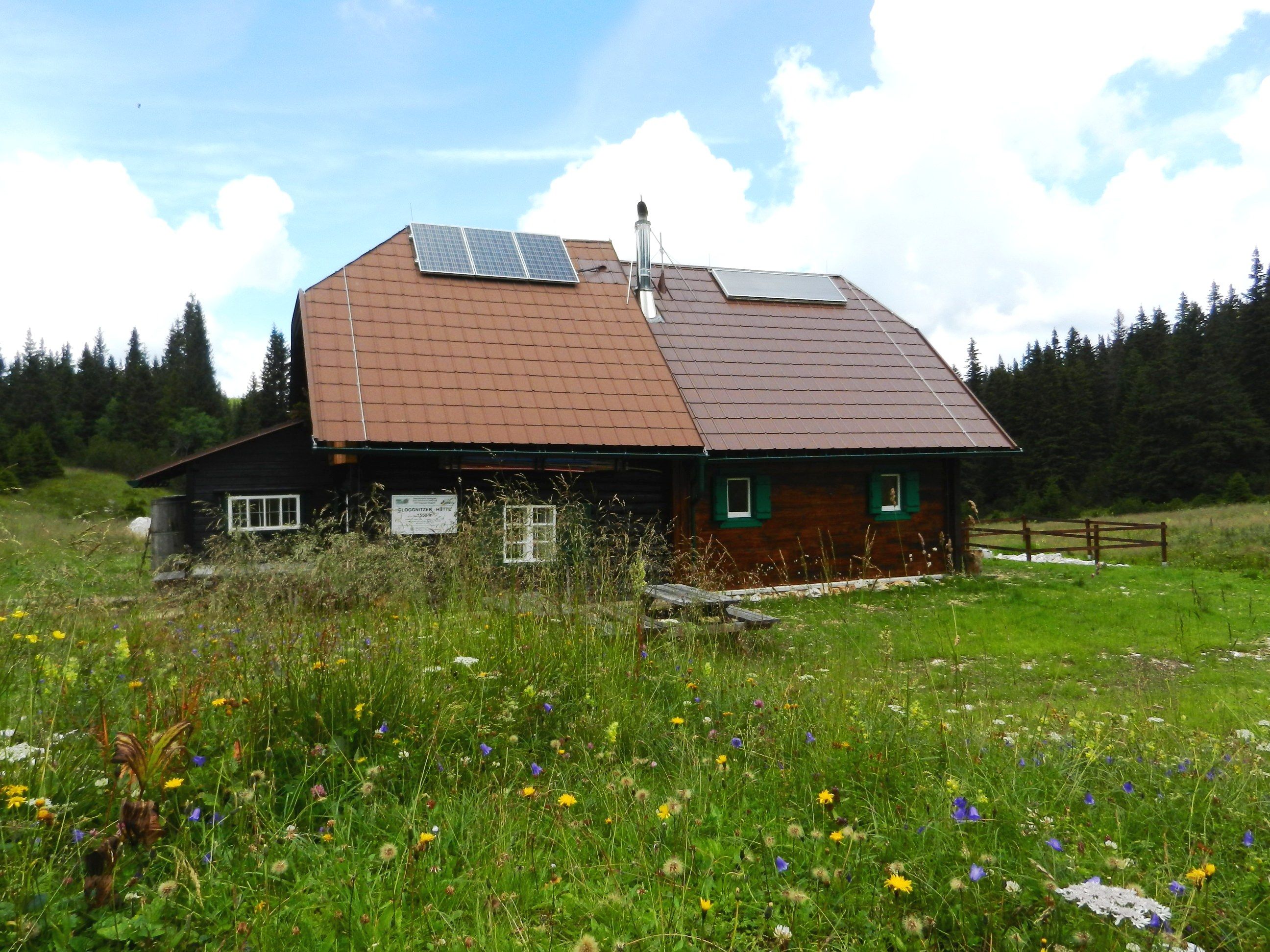 A mountain hut with solar panels on the roof, surrounded by a flowering meadow and forest in the background.