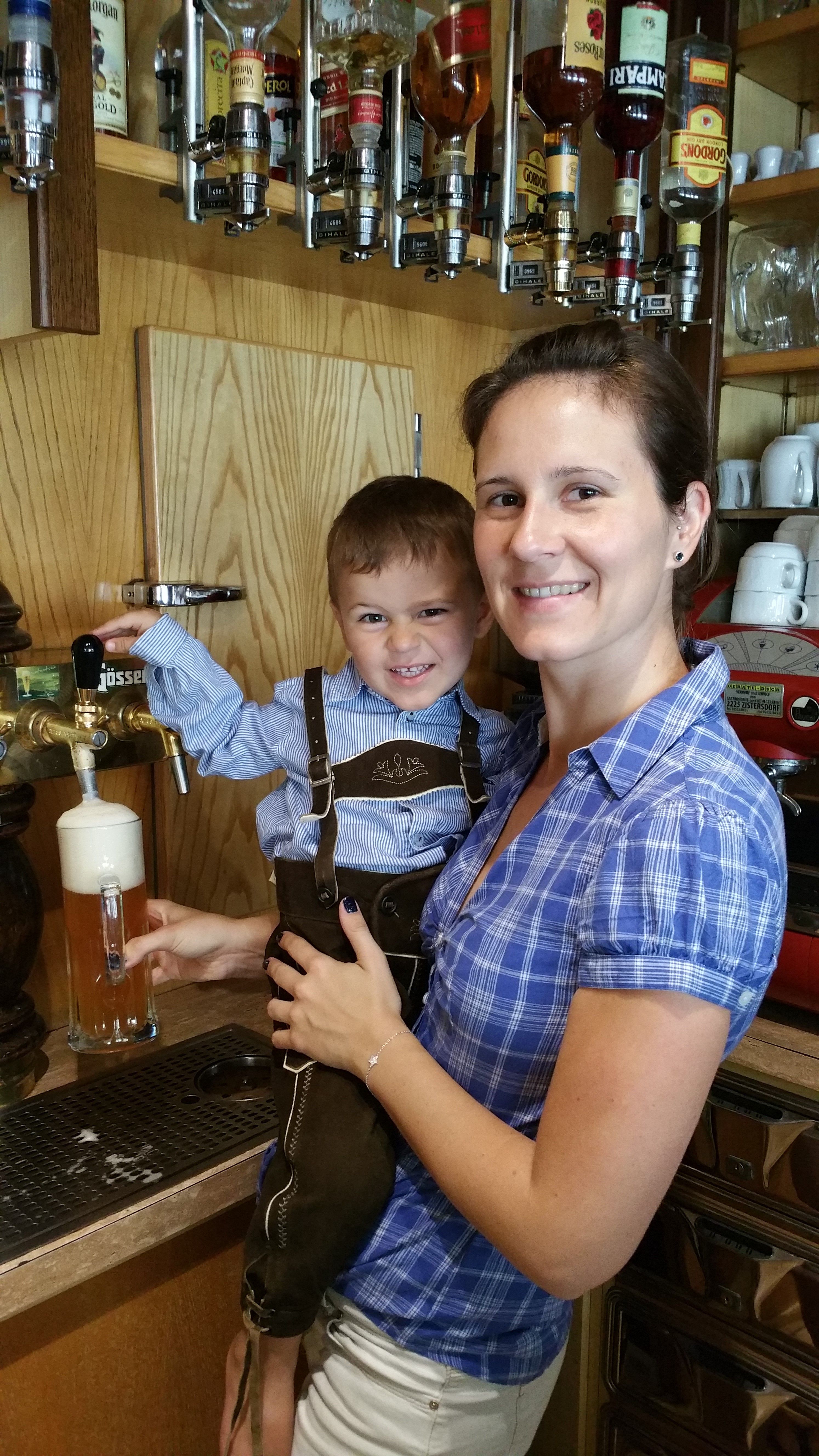 A woman holds a child in lederhosen by a beer tap. Bottles of spirits can be seen in the background.