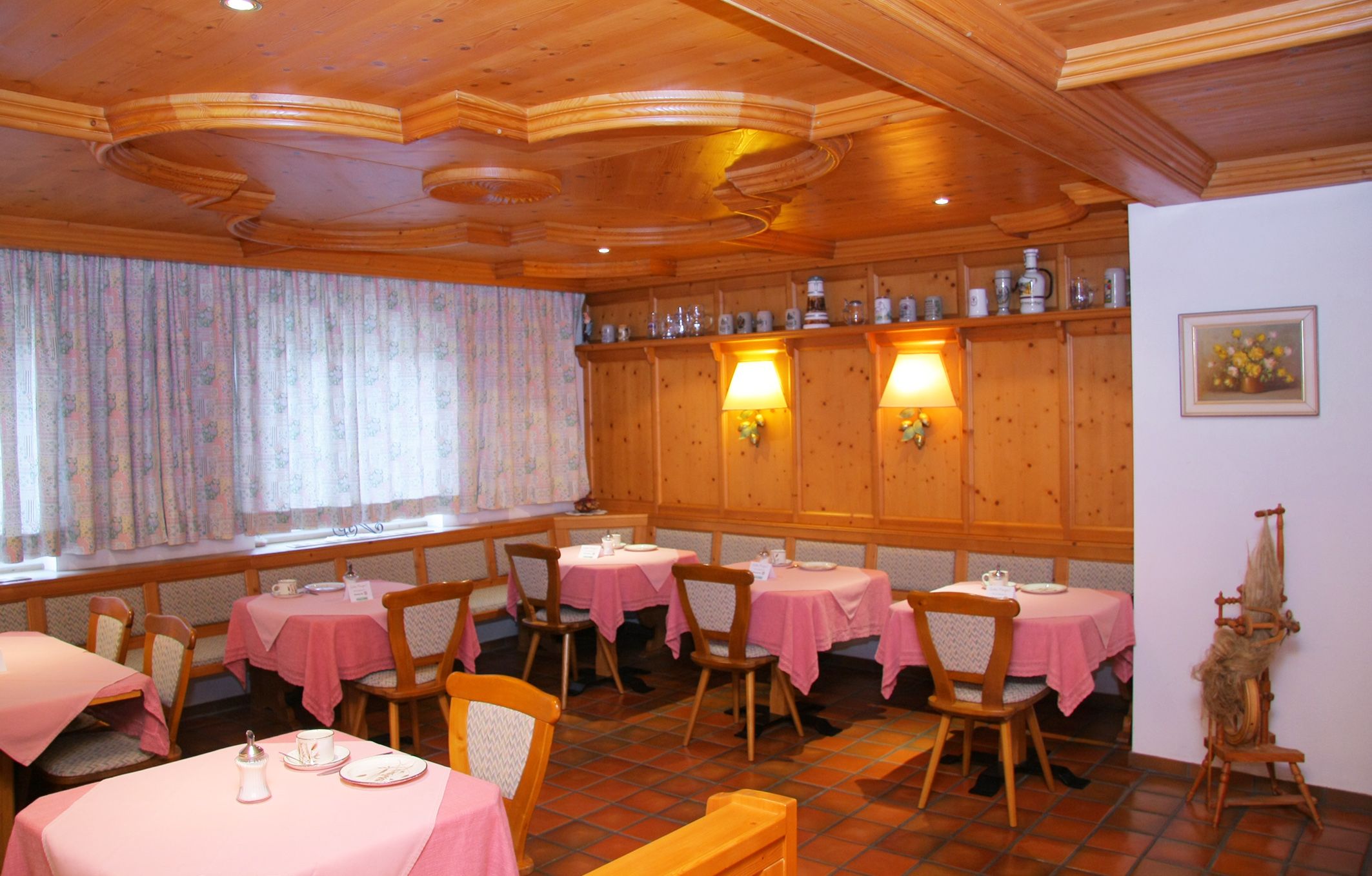 Cozy breakfast room with wooden furniture, pink tablecloths and decorative jugs on a shelf.