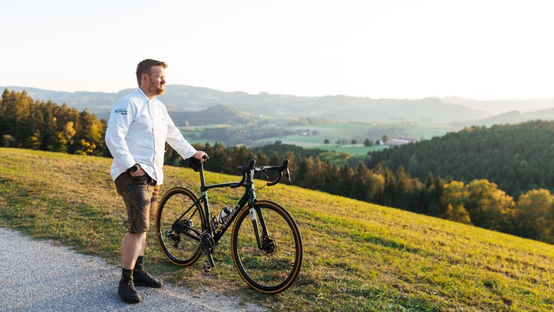 A man in a white shirt stands with a bicycle on a hill overlooking a green landscape.