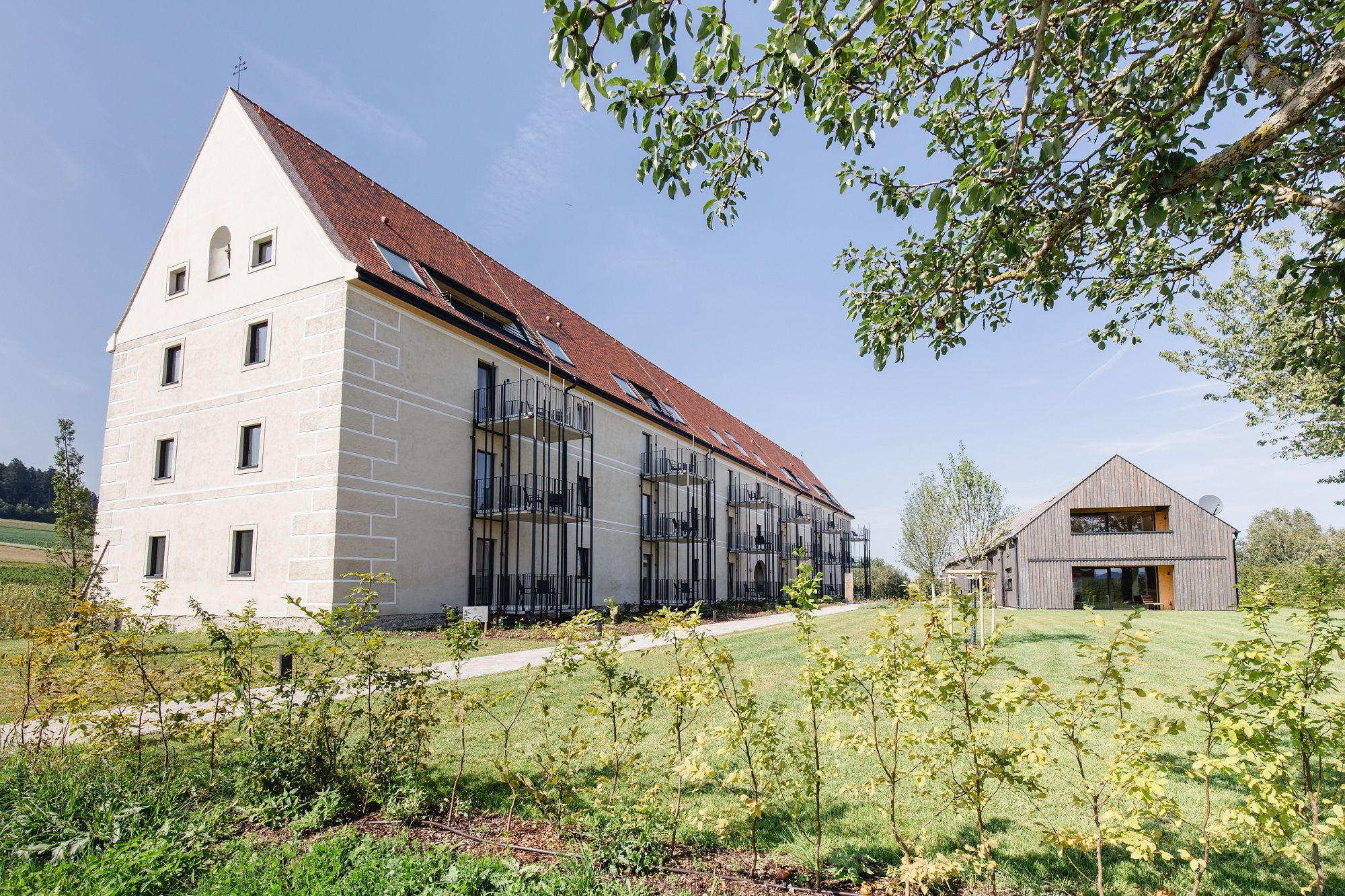 A large, modern building with a red roof and balconies next to a smaller wooden house on a green plot.