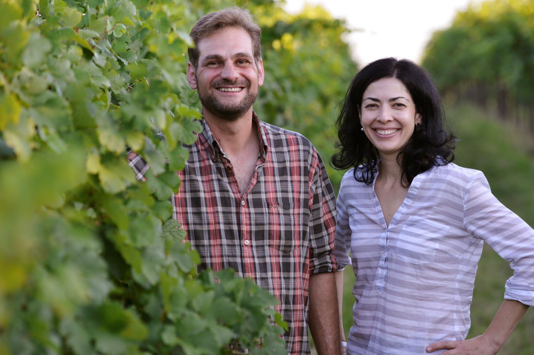 Two people stand smiling in a vineyard.