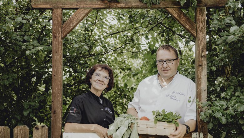 Two people are standing under a wooden arch in the garden, a woman in black clothes and a man in a white shirt with a basket full of vegetables.