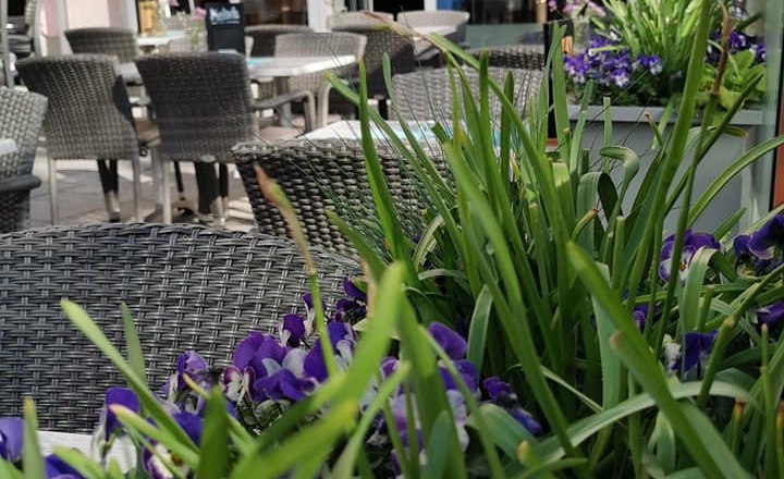 A sidewalk garden with wicker chairs and tables in front of a café, surrounded by blooming flowers and plants.