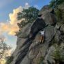 Person climbing on a rock face outdoors, surrounded by trees and blue sky with clouds.