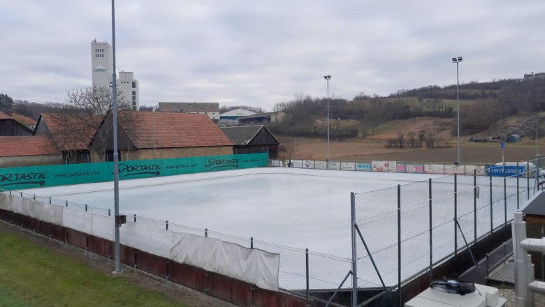 An empty ice rink with surrounding buildings and hills in the background.