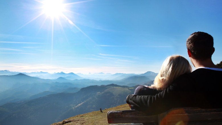 A couple sits on a bench and looks out over a mountain landscape under a bright blue sky and sun.
