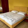 A double bed with a yellow headboard, white comforters and pillows in a room with a yellow wall and red wardrobe.