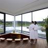 A couple in bathrobes stand in a modern wellness area with a view of a pond.