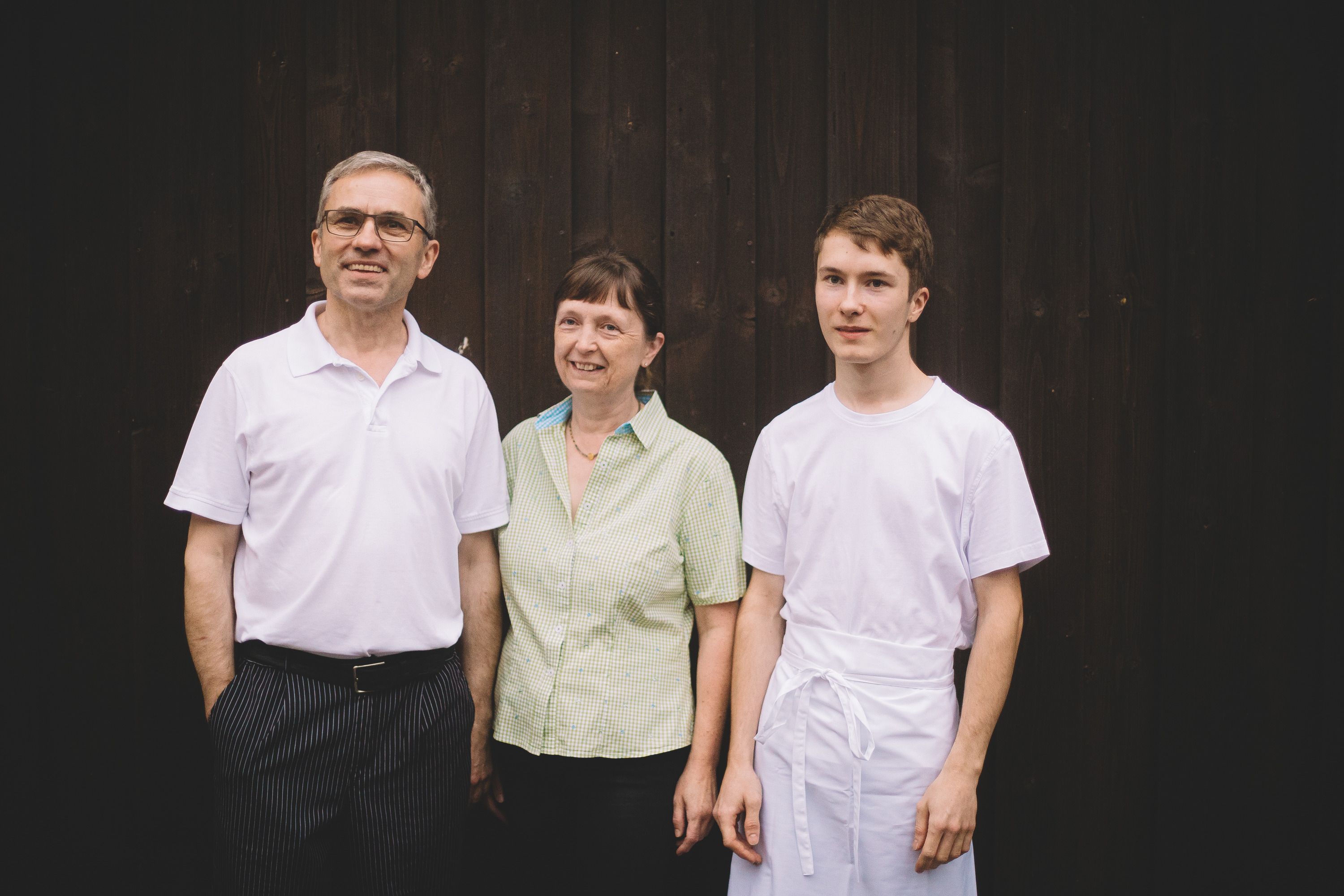 Three people stand in front of a dark wooden wall, smiling and looking into the camera.