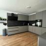 Modern gray kitchen unit with black worktops and wooden floor.