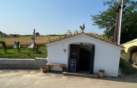 White cellar building with a rooster sculpture on the roof and an open entrance, surrounded by rural landscape.
