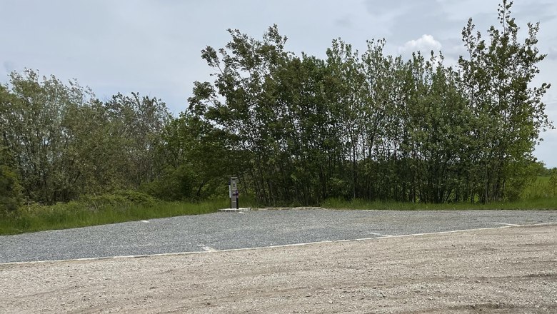 An empty motorhome pitch with a gravel floor, surrounded by trees and a cloudy sky.