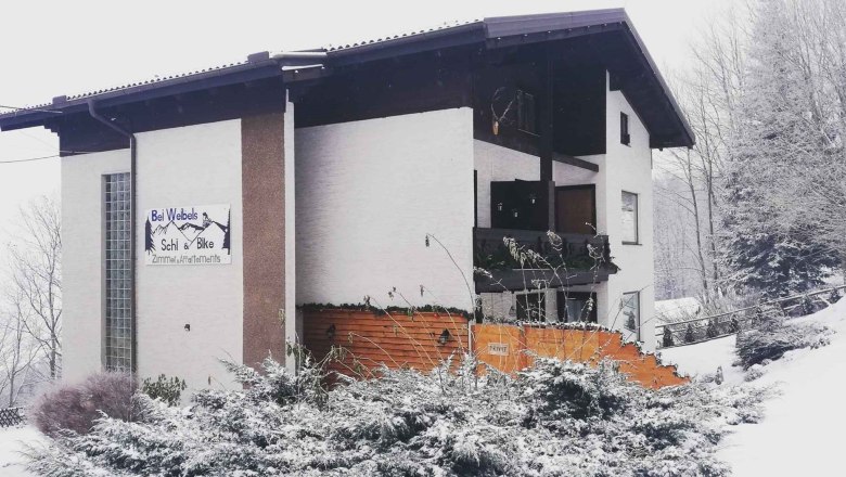 A snow-covered building with a sign 'Bei Webels Ski & Bike Zimmer-Apartments'.